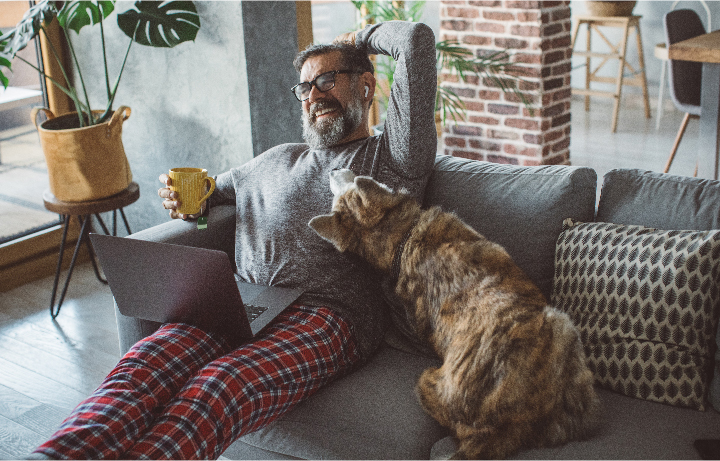 A man holds a cup of coffee as he sits on a couch with his laptop. He laughs as his dog rests its face on his chest